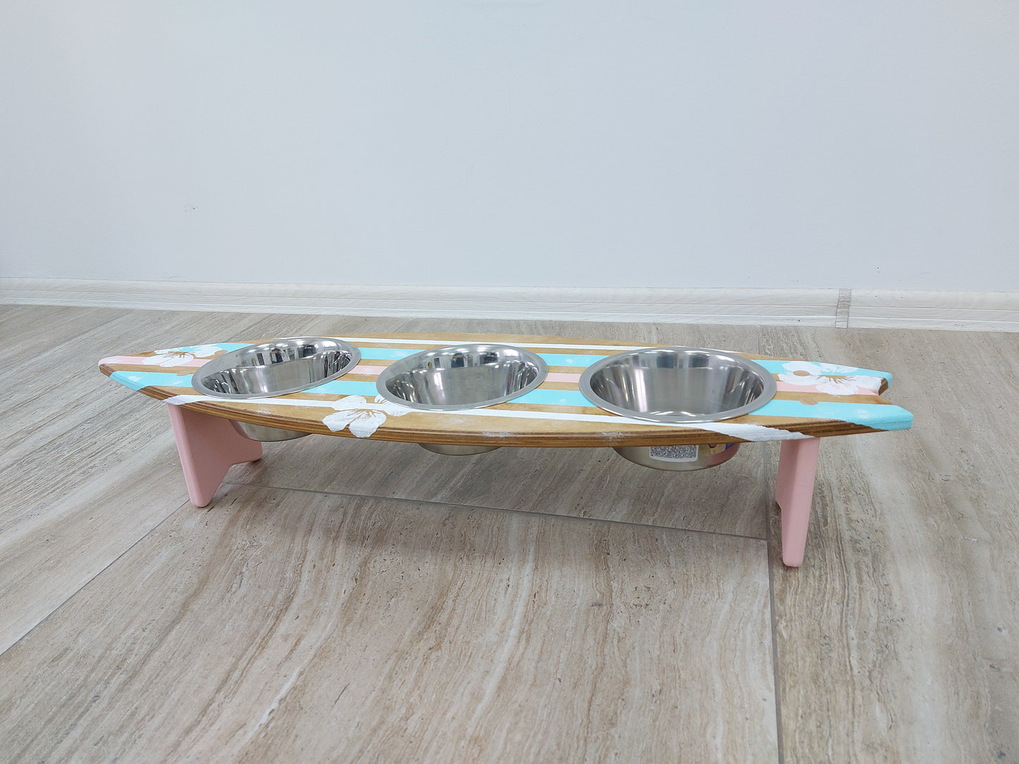Surfboard pet feeding station with three bowls on a wooden floor.