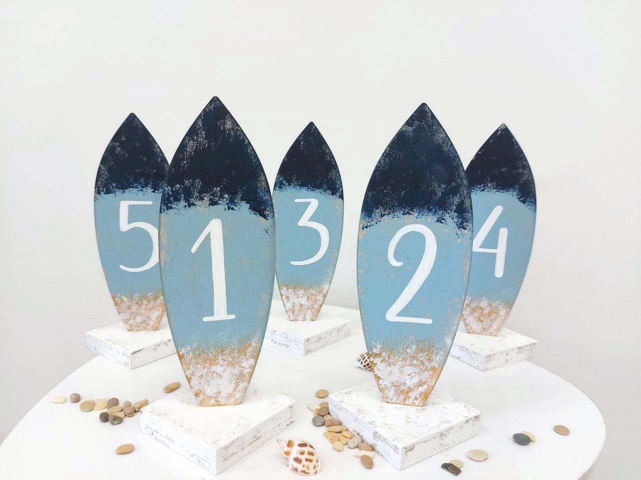 Set of five decorative surfboard-shaped wedding table number signs with sand and water gradient on a white surface.