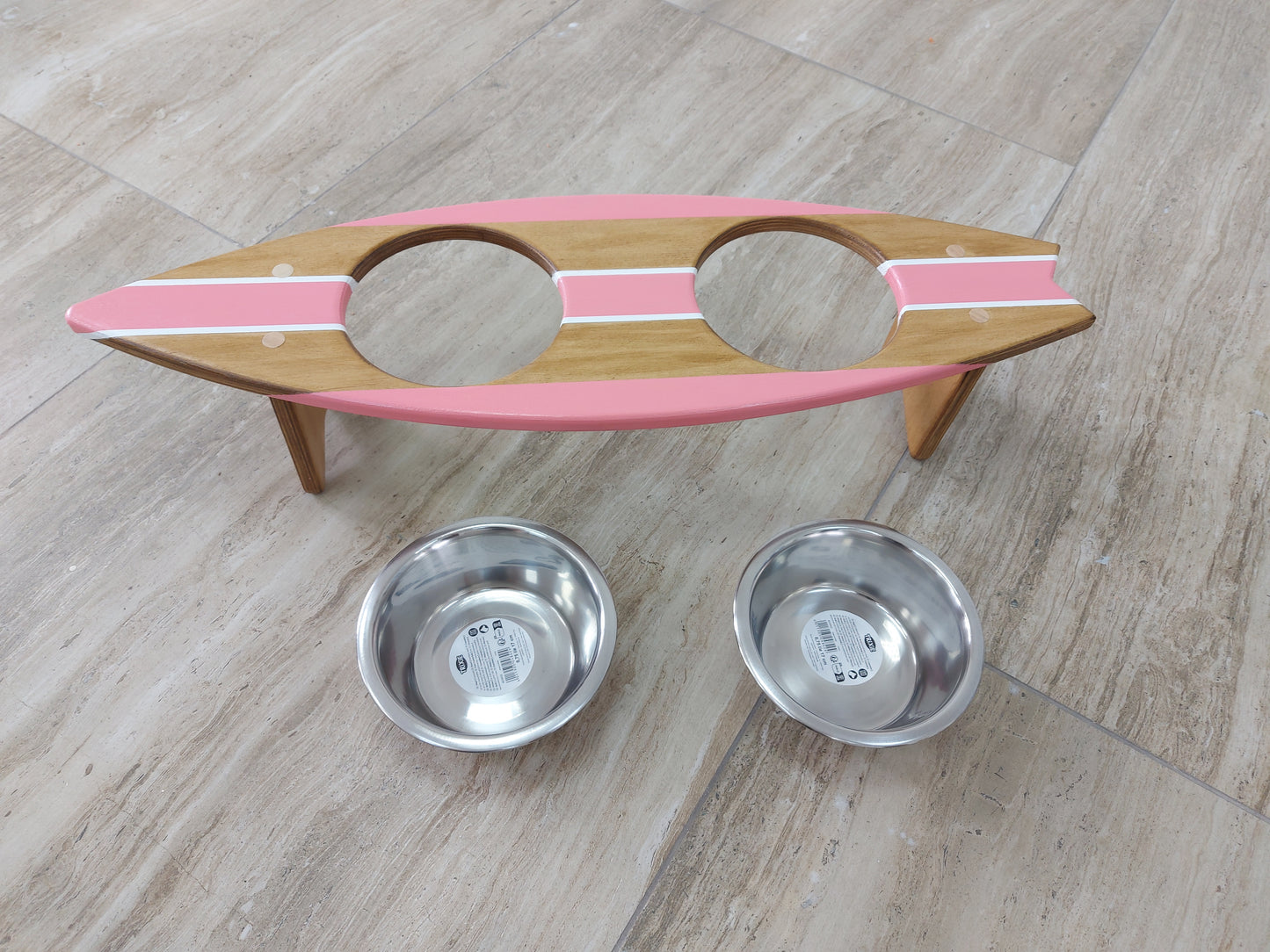 Wooden surfboard pet feeder with pink top and two metal bowls on a wooden floor.