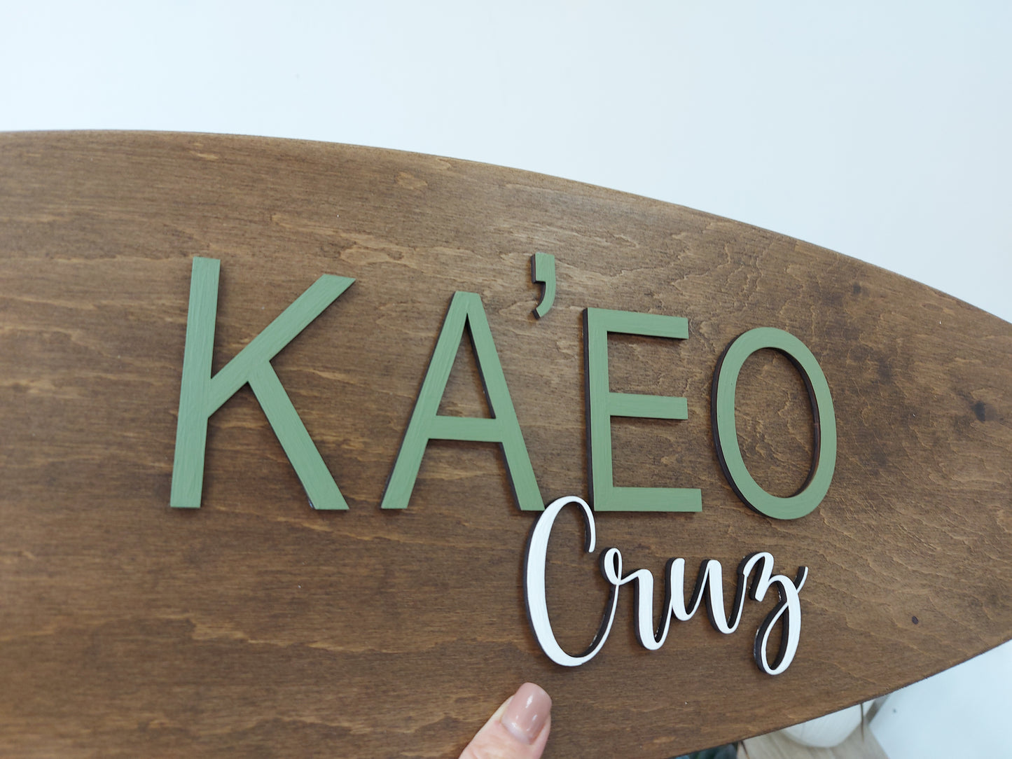 Wooden surfboard-shaped nursery name sign with 'KA'EO Cruz' text on a light blue background