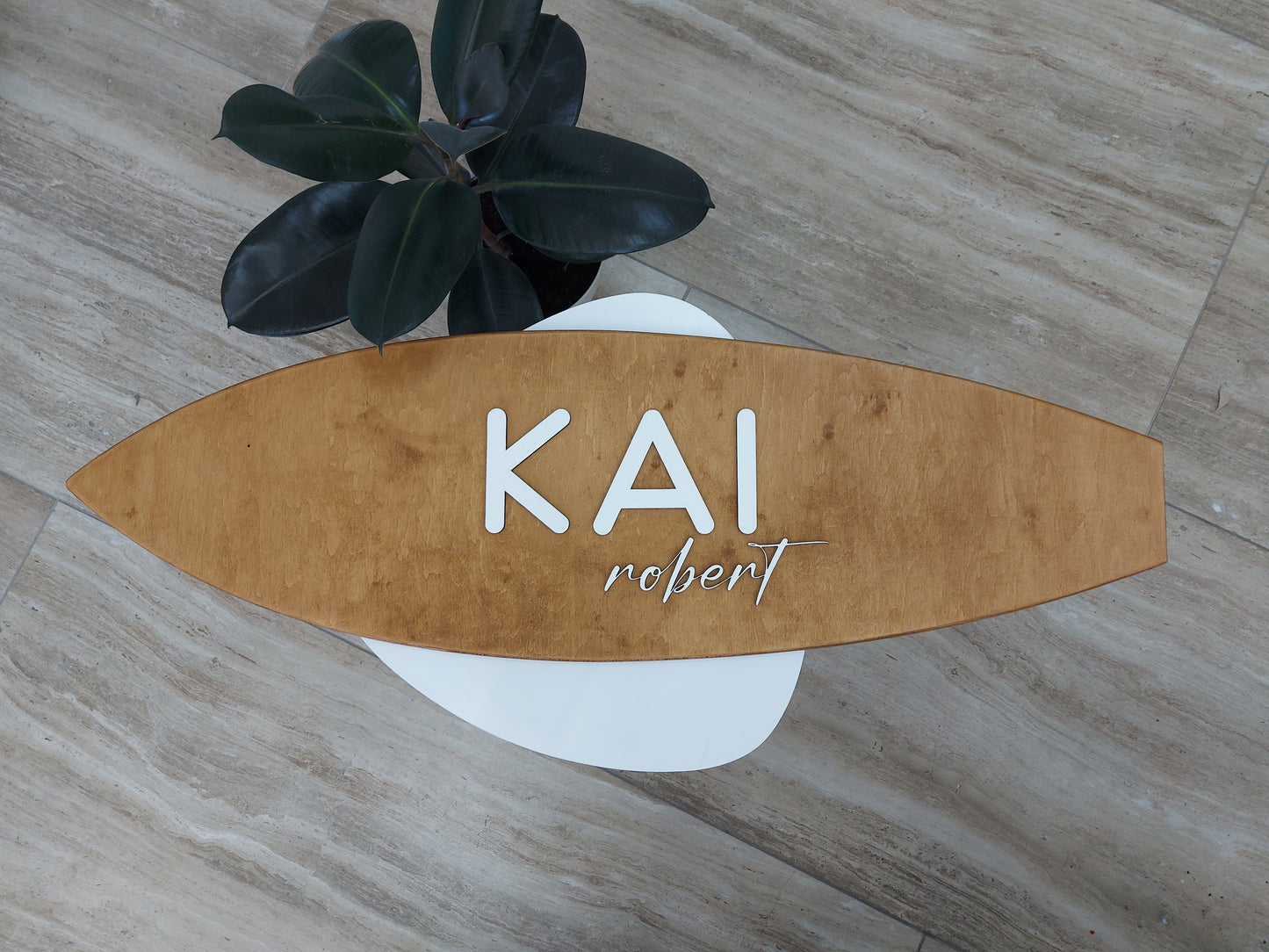 Wooden surfboard-shaped name sign with 'KAI' and 'robert' on a wooden surface.