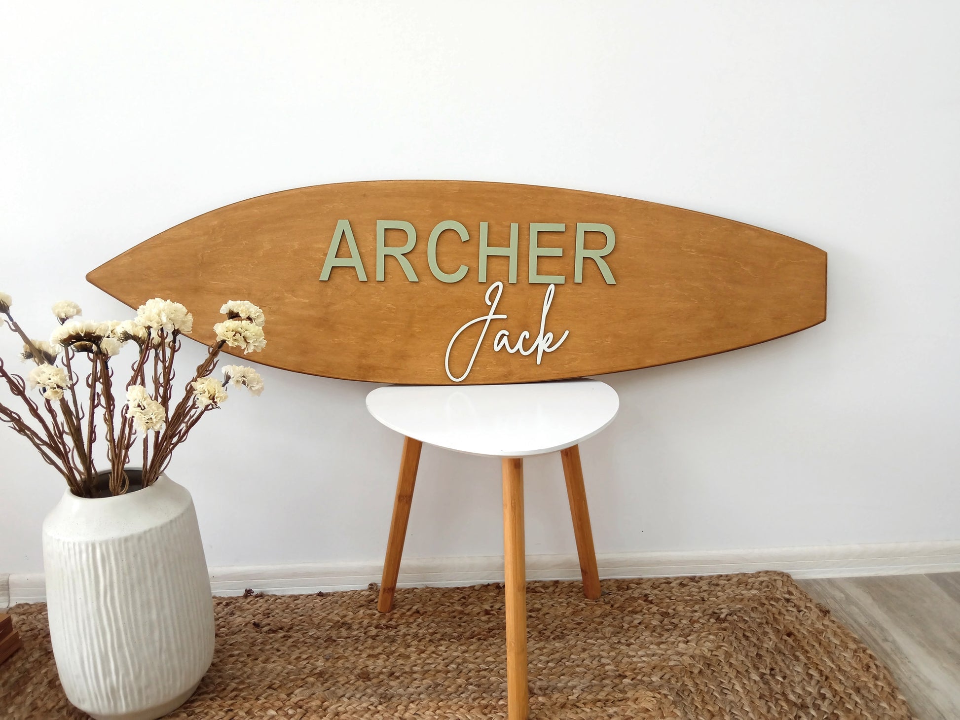 Wooden surfboard-shaped name sign with 'ARCHER Jack' on a white wall with a vase of flowers and a small table.