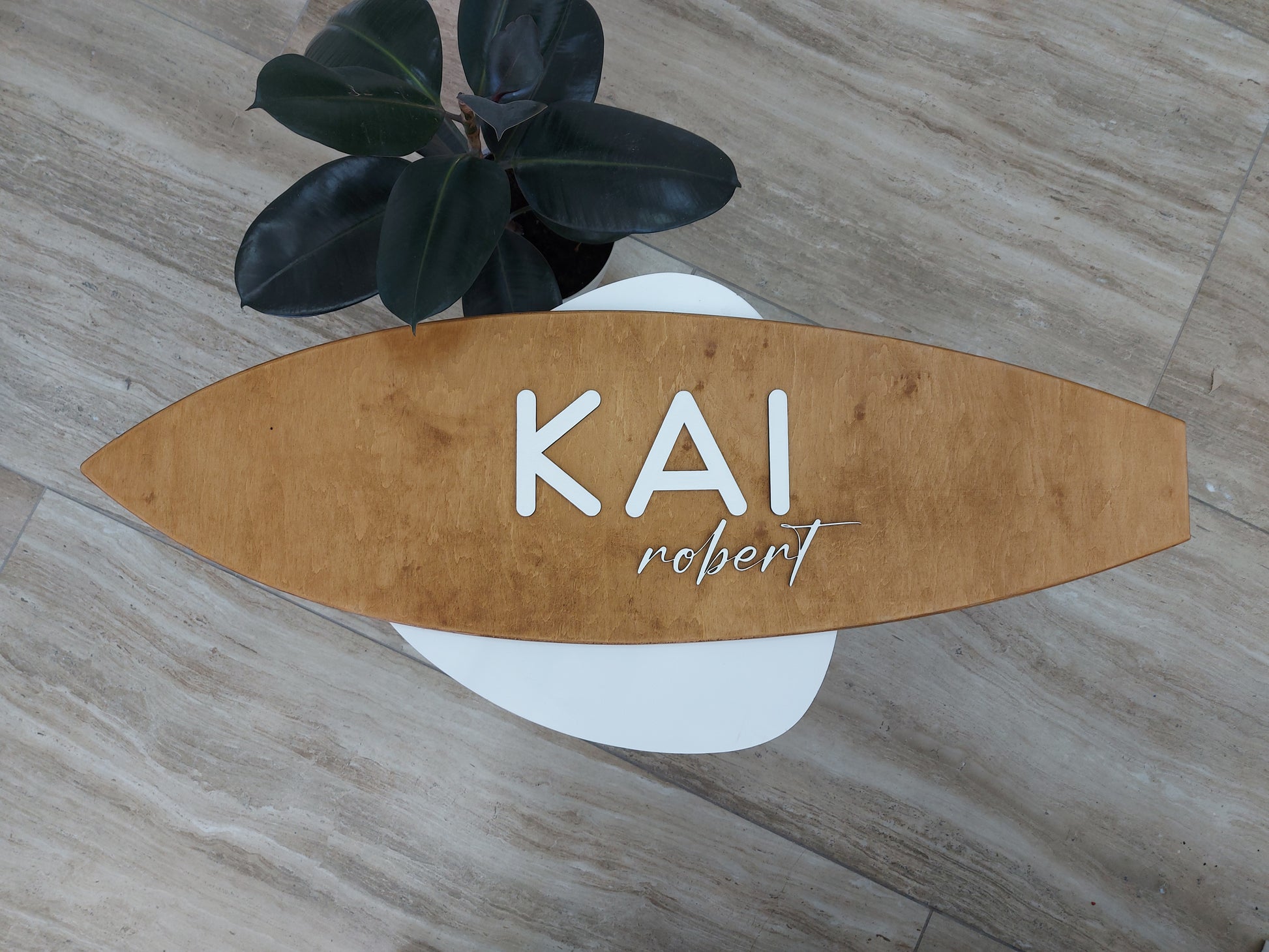 Wooden surfboard-shaped sign with 'KAI' and 'robert' text on a wooden floor.