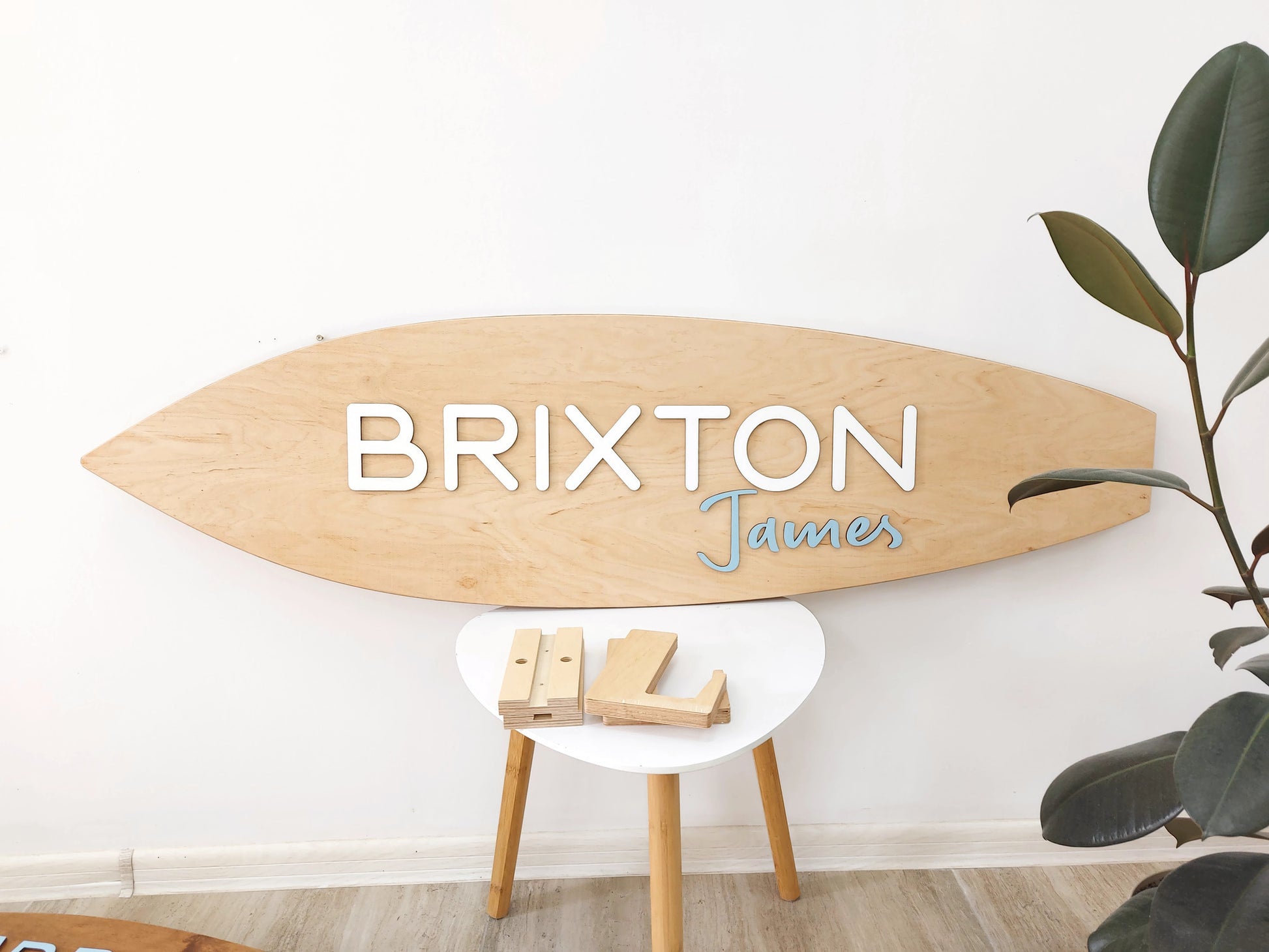 Wooden surfboard-shaped nursery name sign with 'Brixton' and 'James' text on a white wall.