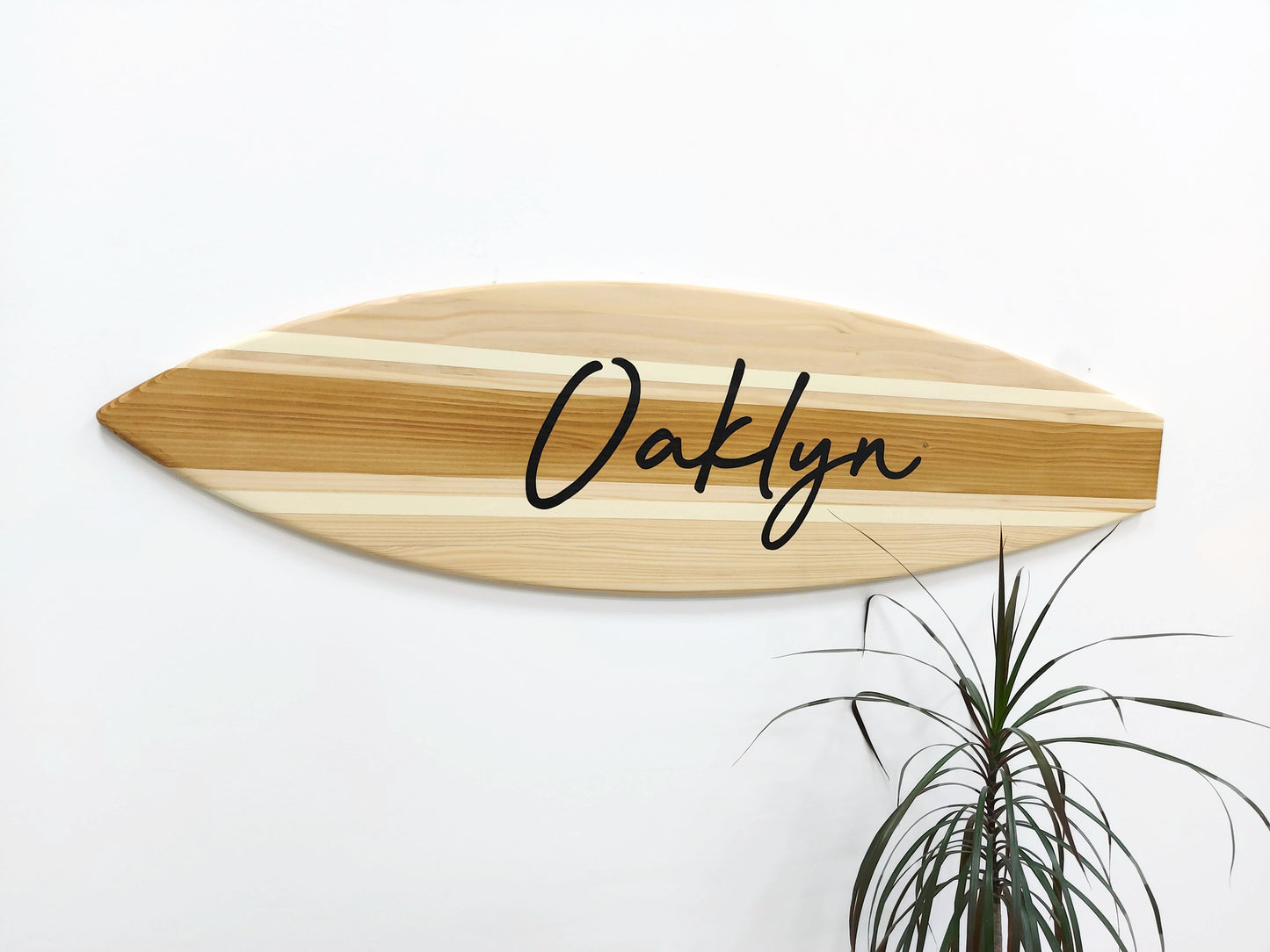 Wooden nameplate with 'Oaklyn' engraved on a white wall with a plant in the corner.