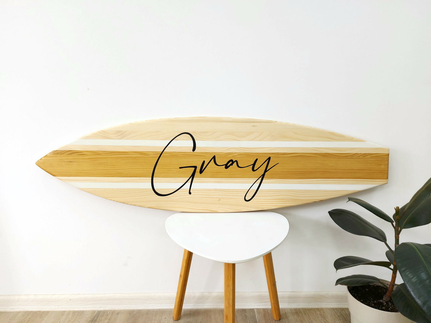 Wooden surfboard-shaped sign with 'Gray' on it, placed on a white stool against a white wall.