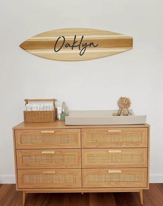 Wooden dresser with wicker drawers, basket, and baby items against a white wall with a wooden surfboard sign.