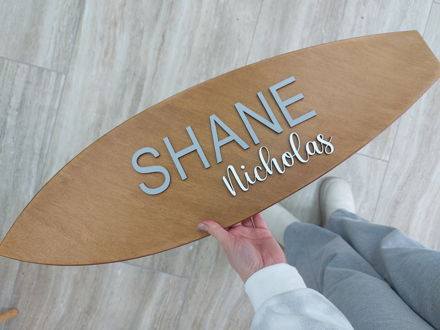 Wooden surfboard-shaped sign with 'SHANE Nicholas' engraved, held by a person on a light wooden floor.