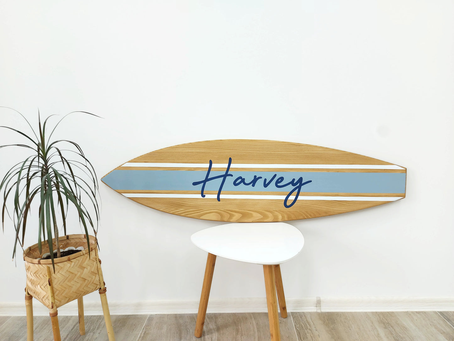 Wooden surfboard name sign with 'Harvey' on it against a white wall, next to a plant and small table.