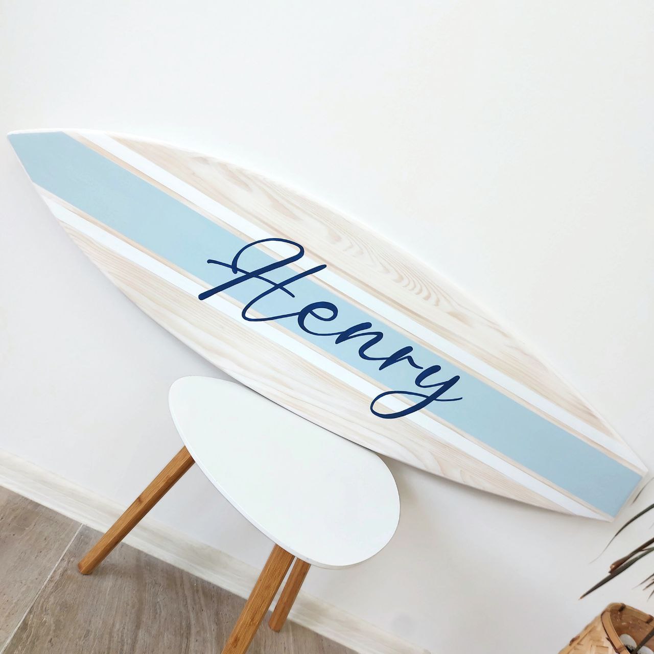 Decorative paddle surfboard name sign with 'Henry' written on it, leaning against a white wall.
