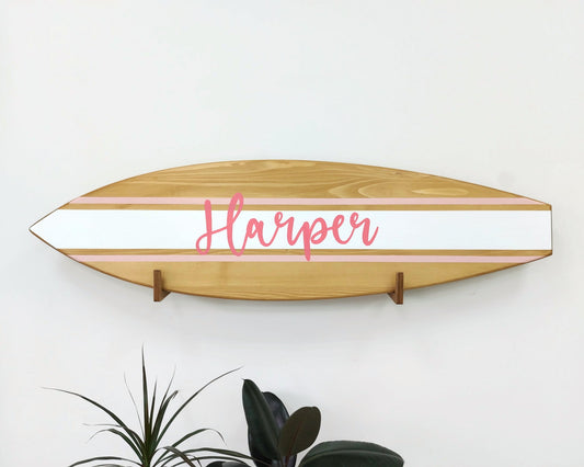 Surfboard-shaped wooden name sign hanging above crib in coastal nursery