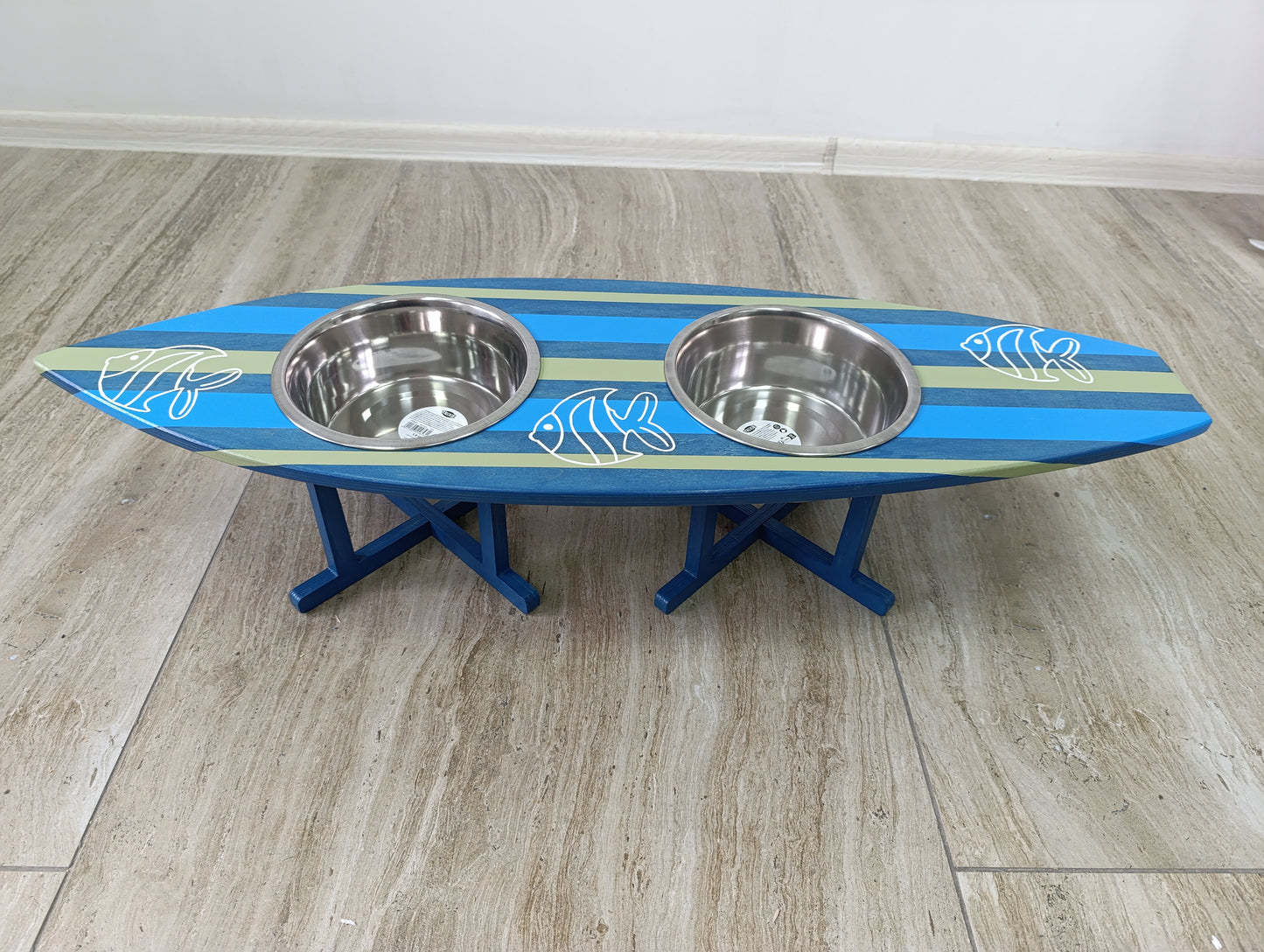 Blue and green surfboard-shaped pet feeder with two metal bowls on a wooden floor.