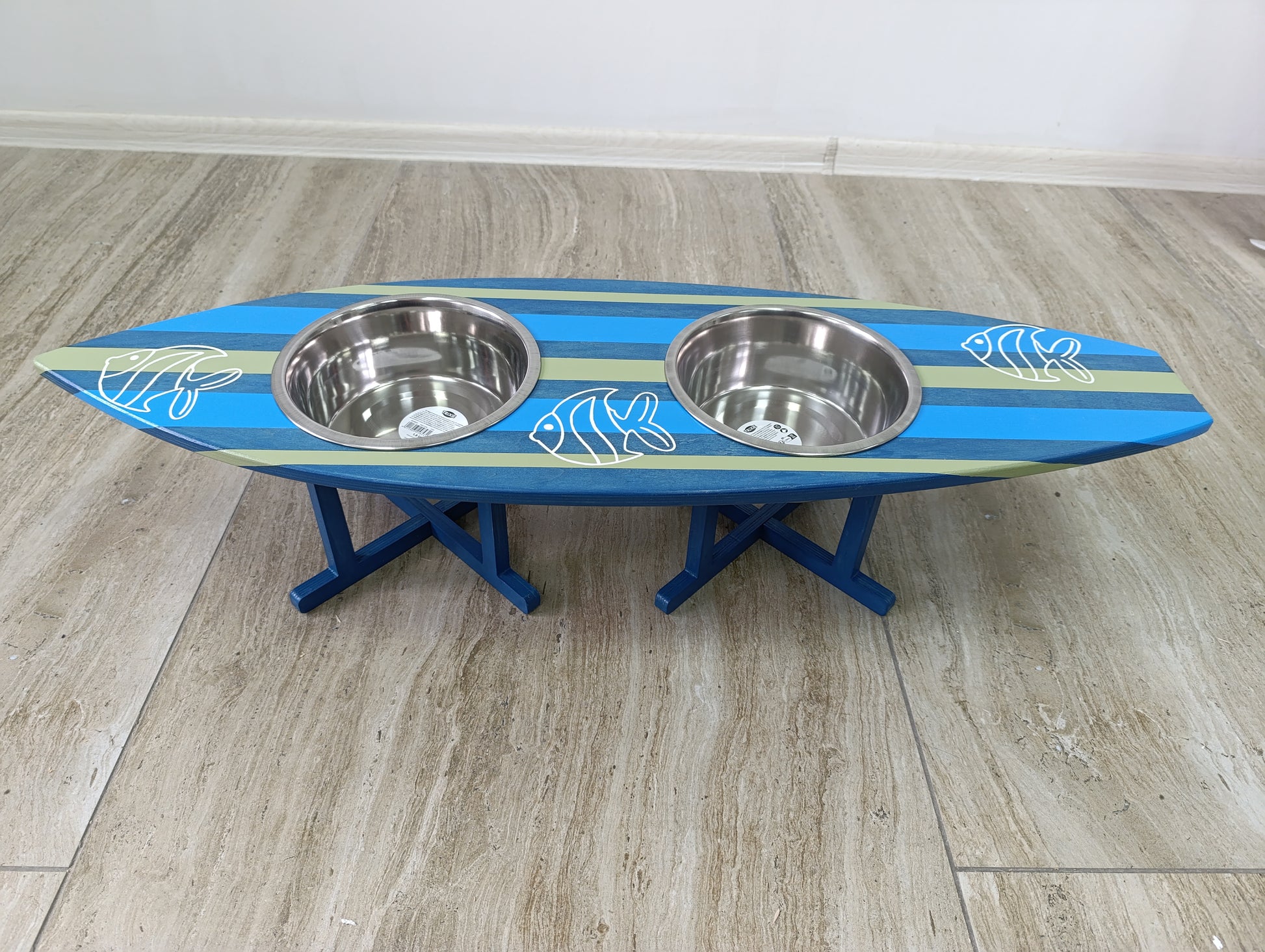 Blue and green surfboard-shaped pet feeder with two metal bowls on a wooden floor.