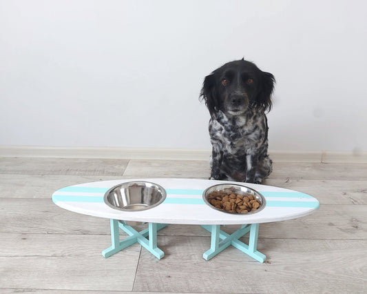 Dog sitting behind a white and blue surfboard pet dining table with two bowls on a wooden floor.
