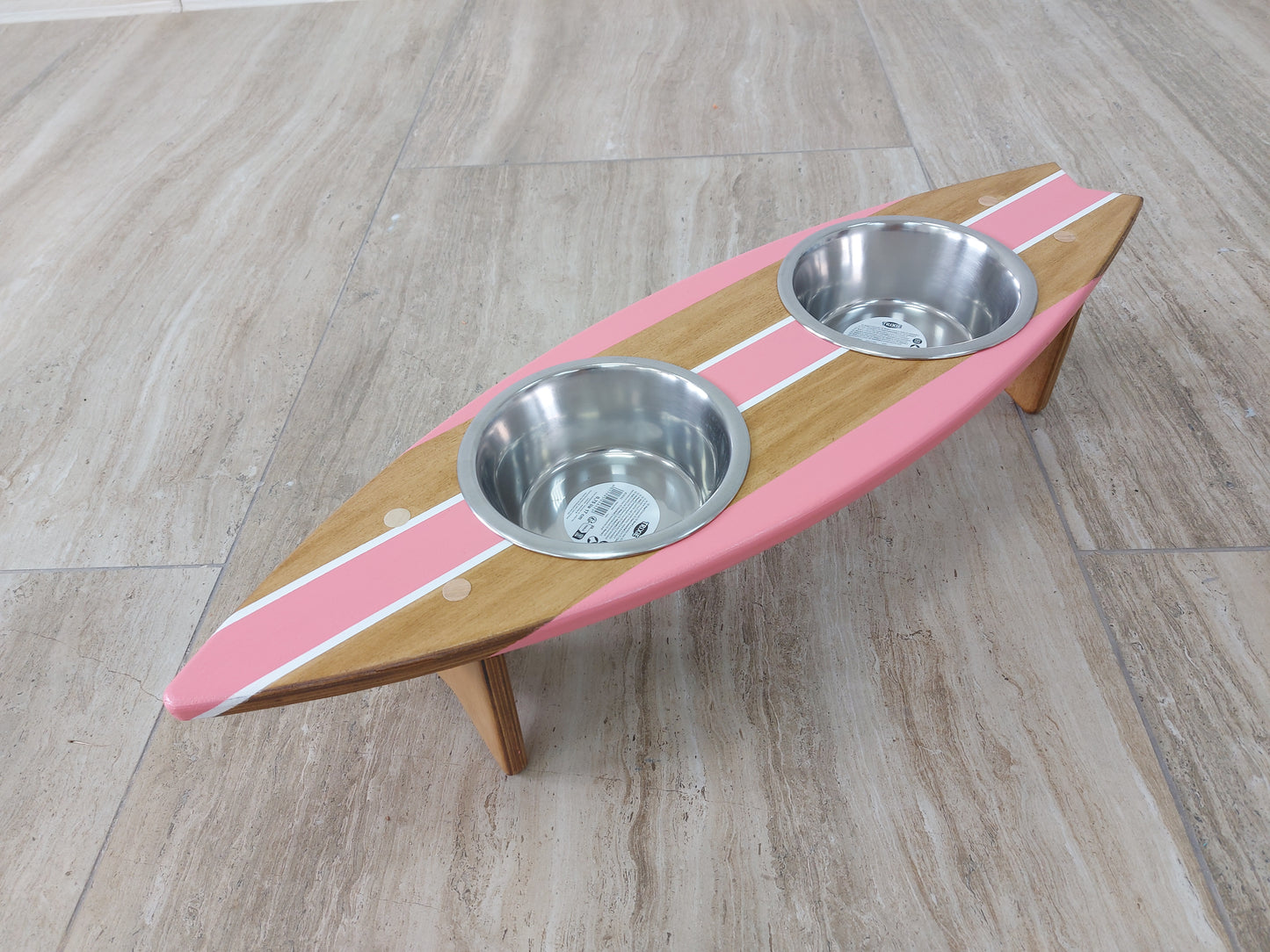 Wooden surfboard pet feeder with two metal bowls on a wooden floor