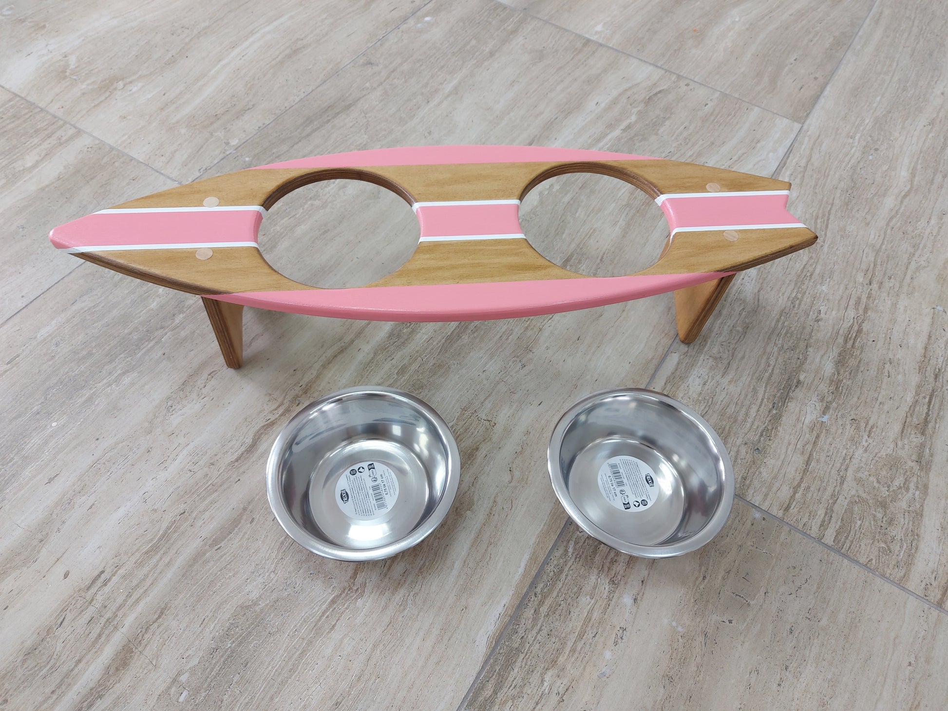 Wooden surfboard pet feeder with pink top and two metal bowls on a wooden floor.