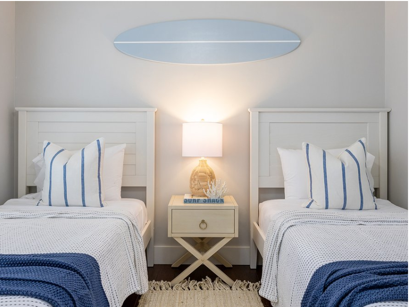 Two twin beds with white and blue bedding in a room with a nightstand , surfboard wall sign decor and lamp.