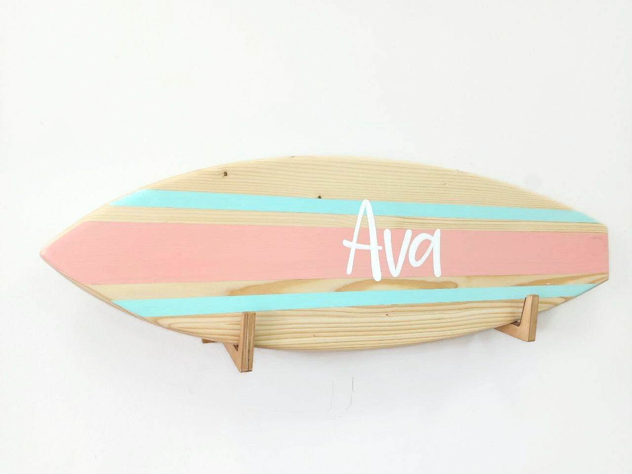 Wooden surfboard-shaped name sign with pastel colors and 'Ava' branding on a white background