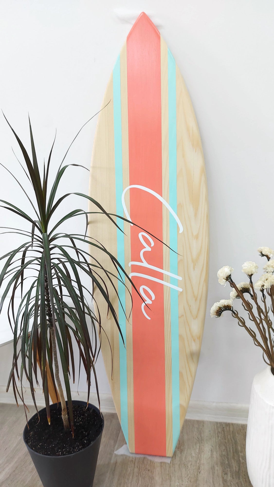 Wooden surfboard name sign with colorful design leaning against a white wall, next to a potted plant and vase.